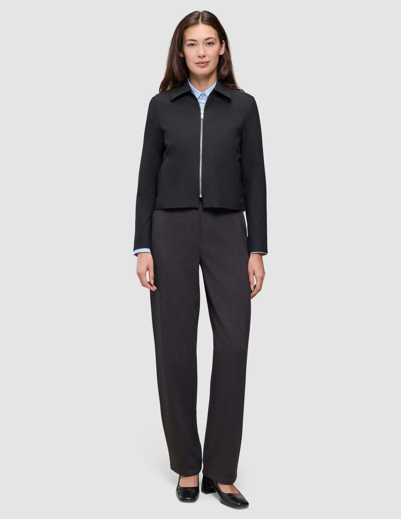 Eine Person trägt eine schwarze Essential Short Zip Jacket über einem hellblauen Hemd mit Knöpfen, dazu eine anthrazitfarbene Hose und schwarze Schuhe mit Blockabsatz.